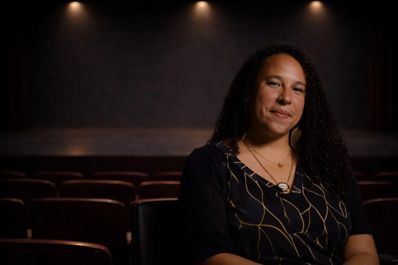 A woman with curly hair and a patterned top sits in a theater auditorium with empty seats and dim lighting in the background.