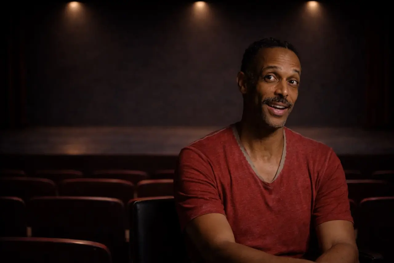 A man in a red shirt sits in a dark, empty theater, smiling slightly, with rows of red seats and a stage in the background.